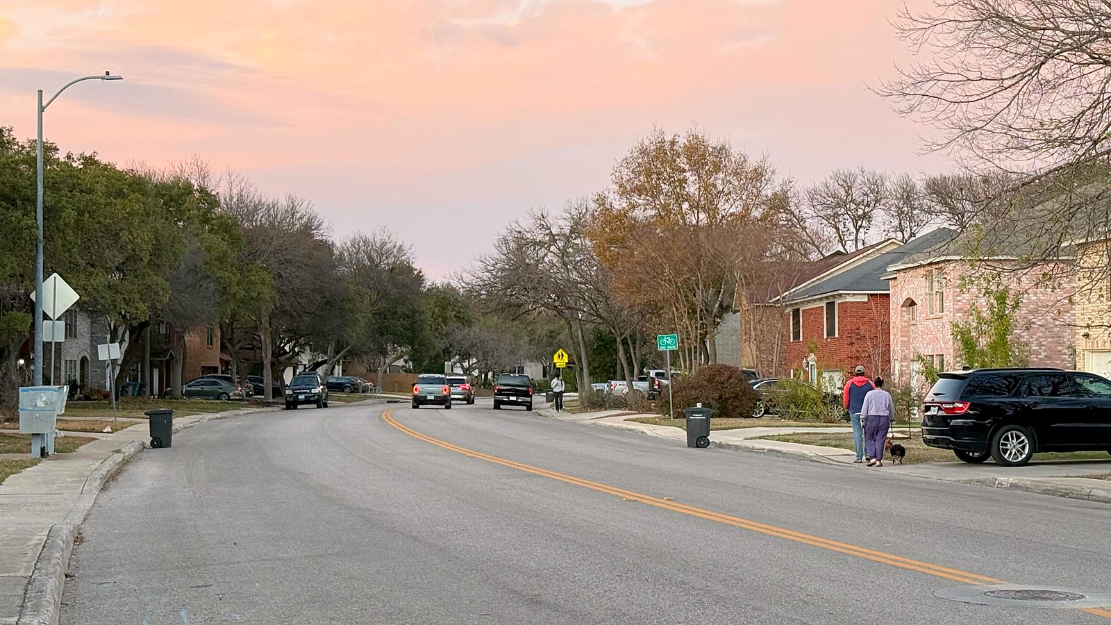 Tree-lined residential street in Parkwood, San Antonio, with neighbors walking a dog at sunset